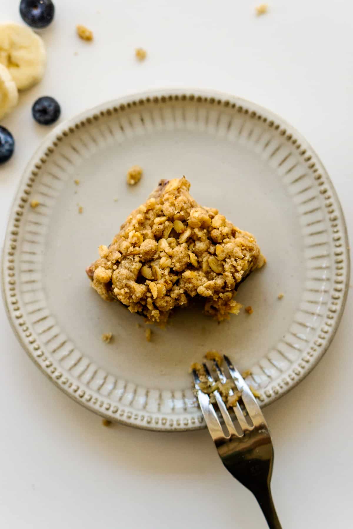 Single banana blueberry oat bar with a bite taken out of it on a white serving plate with a fork placed to the side. 