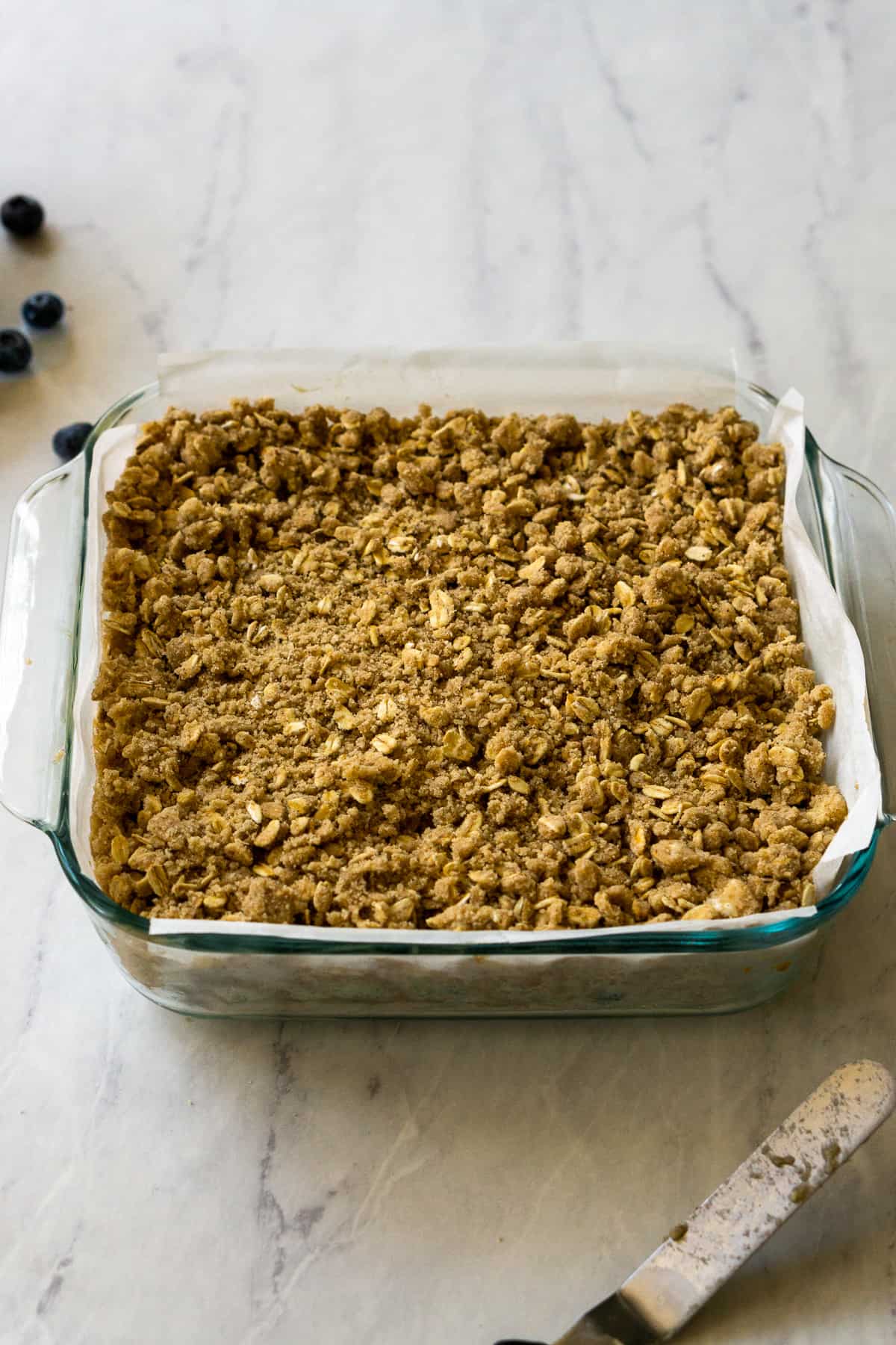 The oat crumble evenly spread across banana blueberry filling and cookie base in an 8x8 inch pan lined with parchment paper. 