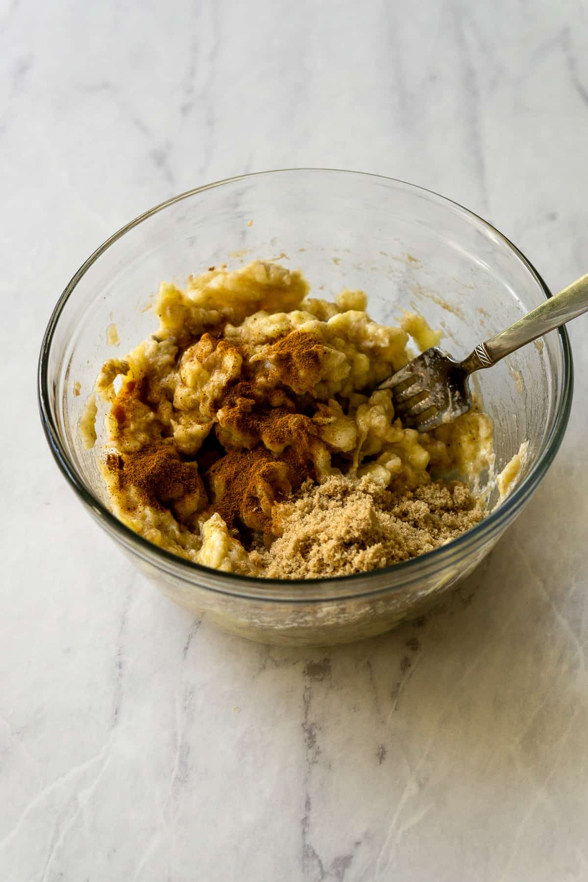 The bananas, brown sugar, and cinnamon being mashed together with a fork in a glass bowl. 